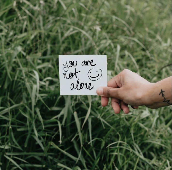 Hand holding a card reading “you are not alone” in a green field